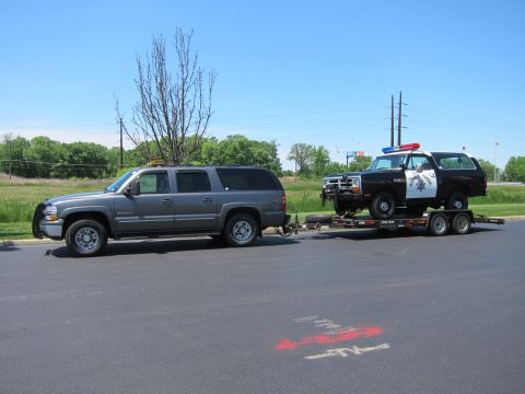 1991 Ford LTD Crown Victoria - gray Chevrolet Suburban tows a flatbed trailer loaded with a black-and-white police SUV across a paved lot.