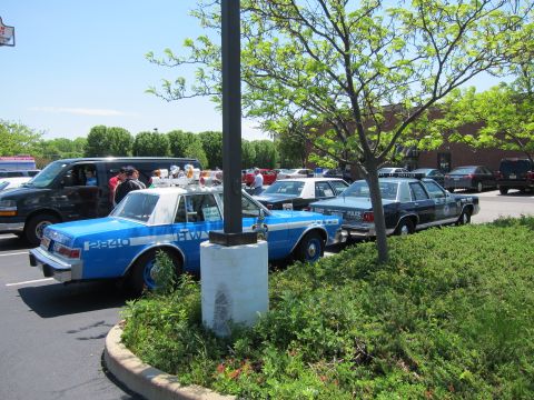 1991 Ford LTD Crown Victoria - blue Highway police sedan marked 2820 and other cruisers parked around a planted lamppost island at a sunny car gathering.
