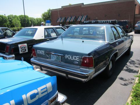 1991 Ford LTD Crown Victoria - Rear three-quarter view of a blue State Police Ford sedan with an Out of Service roof light bar, parked among other cruisers.