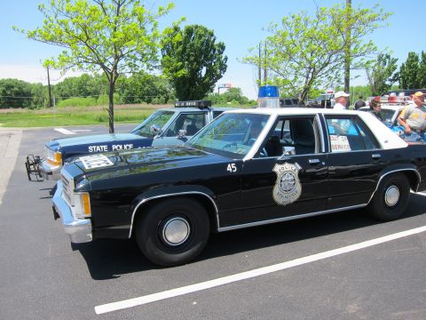 1991 Ford LTD Crown Victoria - black-and-white Police Ford sedan marked 45 with a gold badge decal and roof light bar parked at a sunny car gathering.