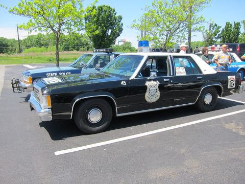1991 Ford LTD Crown Victoria - black-and-white Police Ford sedan marked 45 with gold badge decal and light bar parked beside a blue State Police car.