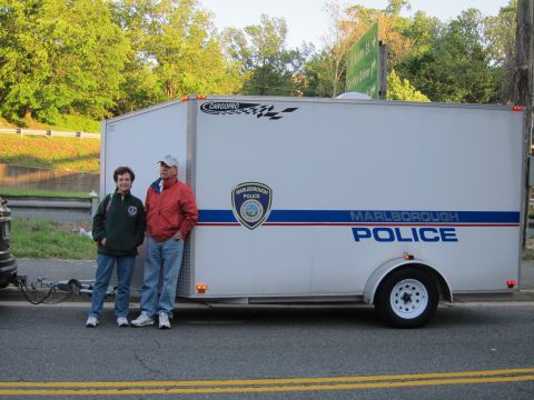 1991 Ford LTD Crown Victoria - Two people pose beside a white Marlborough Police enclosed cargo trailer with badge and blue stripe at the roadside.