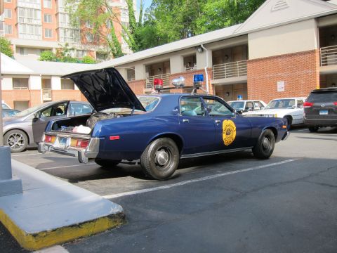 1991 Ford LTD Crown Victoria - dark blue 1970s police sedan parked with its trunk open, displaying a gold star-shaped door emblem in a brick-building parking lot.
