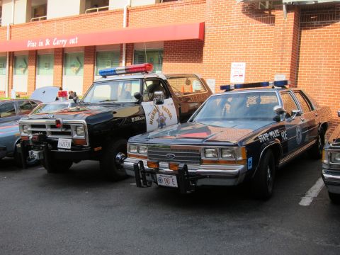 1991 Ford LTD Crown Victoria - black Ford state-police sedan with lightbar parked next to a black Dodge state-police pickup outside a brick restaurant.