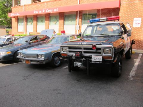 1991 Ford LTD Crown Victoria - black Dodge police pickup fitted with a front winch parked beside vintage patrol sedans outside a brick Dine in & Carry out eatery.