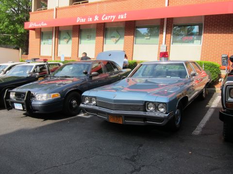 1991 Ford LTD Crown Victoria - light blue 1970s police sedan with its trunk open parked next to a dark Ford patrol car outside a brick restaurant.