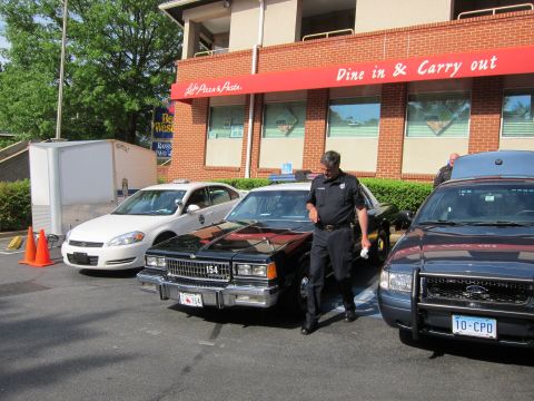 1991 Ford LTD Crown Victoria - uniformed officer walks past a lineup of police cars including a white Chevrolet, a black sedan numbered 154, and a dark patrol car.
