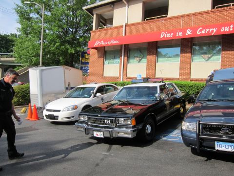 1991 Ford LTD Crown Victoria - man approaches a row of patrol cars, including a white Chevrolet and a black sedan marked 154, outside a brick Dine in & Carry out restaura…