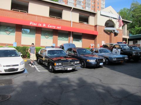 1991 Ford LTD Crown Victoria - man stands beside a row of black police cars including a sedan marked 154, a Ford patrol car, and a vintage truck under an American flag.