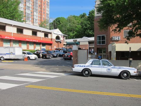 1991 Ford LTD Crown Victoria - white-and-blue police sedan parked on the street in front of a brick building numbered 1501, with more patrol cars in a lot beyond.