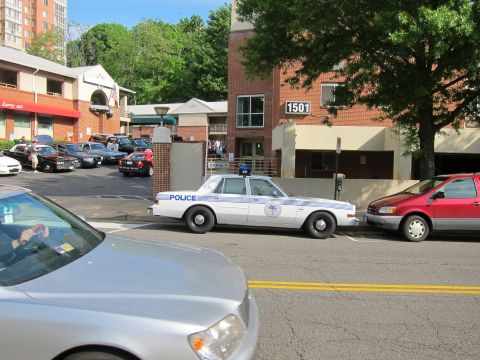 1991 Ford LTD Crown Victoria - white police patrol sedan parked at the curb outside a brick building numbered 1501, with a silver car passing in the foreground.