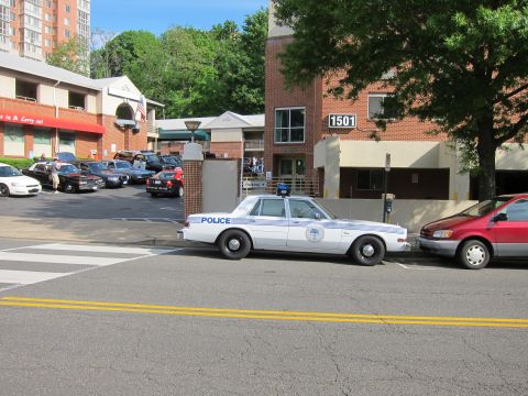 1991 Ford LTD Crown Victoria - white police sedan parked at a crosswalk in front of a brick building numbered 1501, with a red minivan and more cars nearby.