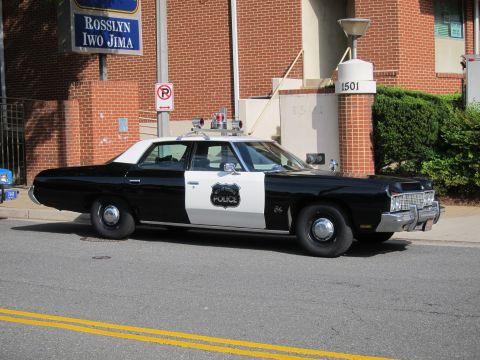 1991 Ford LTD Crown Victoria - black-and-white vintage police sedan with a roof lightbar and door shield parked along a curb near a brick building marked 1501.
