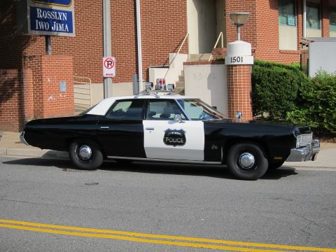 1991 Ford LTD Crown Victoria - black-and-white vintage police sedan with door shield and roof lightbar parked at the curb beside a Rosslyn Iwo Jima sign.
