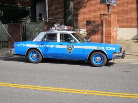 1991 Ford LTD Crown Victoria - blue NYPD police sedan with white doors, roof lightbar, and HWY markings parked at the curb against a brick wall.
