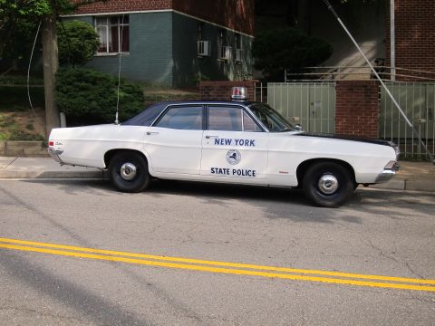 1991 Ford LTD Crown Victoria - Side profile of a white New York State Police sedan with door seal and roof light parked along a curb by a building.