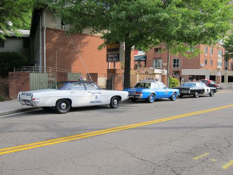 1991 Ford LTD Crown Victoria - lineup of vintage patrol cars led by a white New York State Police sedan, parked along a street by brick buildings and a yellow centerline.