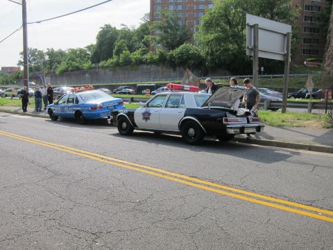1991 Ford LTD Crown Victoria - Several people gather around a black-and-white police sedan with its trunk open at the roadside, with a blue patrol car ahead and parking l…