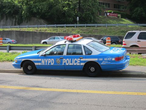 1991 Ford LTD Crown Victoria - blue-and-white NYPD police sedan marked 103 PCT parked at the curb beside a guardrail, with a grassy slope and highway behind.