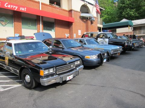 1991 Ford LTD Crown Victoria - lineup of vintage patrol cars including a black sedan marked 154 and a dark Ford, angle-parked outside a brick building flying an American…