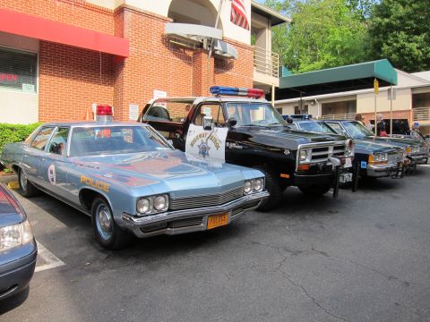 1991 Ford LTD Crown Victoria - light blue police station wagon and a black highway-patrol pickup parked among other vintage patrol cars outside a brick building.