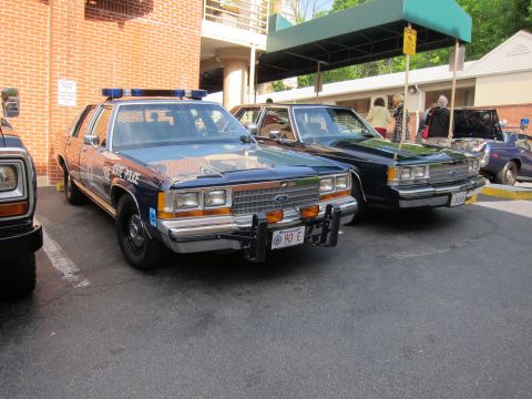 1991 Ford LTD Crown Victoria - Two dark blue Ford state-police sedans with roof lightbars angle-parked side by side in a lot near a building with a green awning.