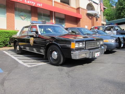 1991 Ford LTD Crown Victoria - black Chevrolet police sedan marked 154 with a roof lightbar angle-parked outside a brick restaurant beside other patrol cars and a US flag.