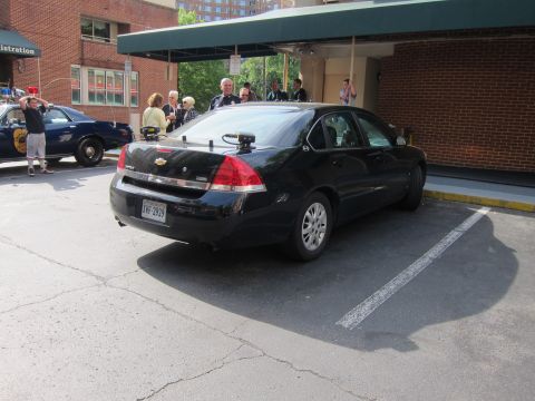 1991 Ford LTD Crown Victoria - black Chevrolet Impala unmarked police sedan with a pillar spotlight sits in a parking lot beside a brick building as people gather nearby.