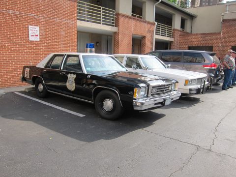 1991 Ford LTD Crown Victoria - vintage black-and-white police cruiser with a star badge decal and roof light sits parked along a brick building beside other cars.