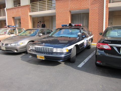 1991 Ford LTD Crown Victoria - dark blue and white Chevrolet Caprice police cruiser with a roof lightbar sits between a tan sedan and a black Toyota Corolla outside a bri…