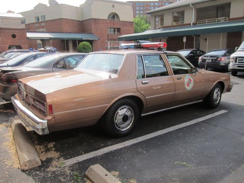 1991 Ford LTD Crown Victoria - tan Chevrolet sedan with a roof lightbar and circular door emblem sits parked outside a hotel conference center among other cars.