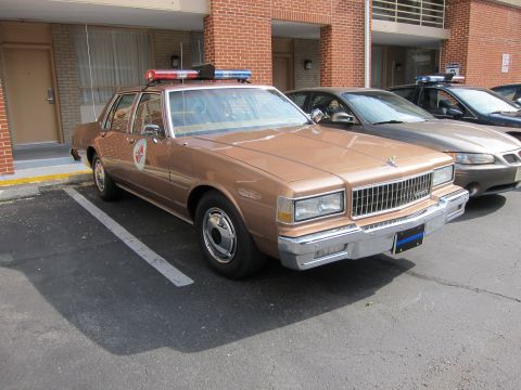 1991 Ford LTD Crown Victoria - tan Chevrolet Caprice police sedan with a roof lightbar and star door emblem sits parked beside a brick building and another sedan.