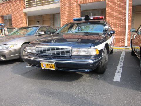 1991 Ford LTD Crown Victoria - dark blue and white Chevrolet Caprice police cruiser with a roof lightbar sits parked in a lot beside a brick building and other cars.
