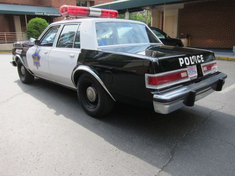 1991 Ford LTD Crown Victoria - black-and-white police sedan marked POLICE with a roof lightbar and star emblem sits parked outside a brick guest registration building.