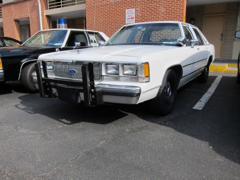 1991 Ford LTD Crown Victoria - white Ford LTD Crown Victoria police sedan fitted with a front push bar sits parked beside a black-and-white cruiser along a brick building.