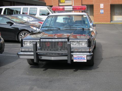 1991 Ford LTD Crown Victoria - black police sedan with a front push bar, roof lightbar, and a POLICE MEMORIAL license plate sits parked in a lot among other cars.