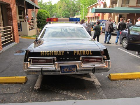 1991 Ford LTD Crown Victoria - black Dodge marked HIGHWAY PATROL with a roof lightbar sits parked outside a motel as a group of people stand nearby.