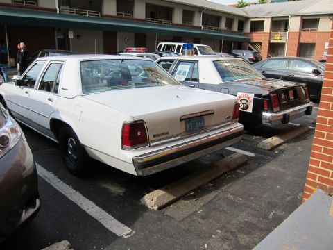 1991 Ford LTD Crown Victoria - white Ford Crown Victoria police sedan sits parked among other cruisers in a motel parking lot beside a brick wall.