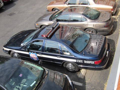 1991 Ford LTD Crown Victoria - overhead view of a dark blue Chevrolet Caprice marked STATE TROOPER with a roof lightbar parked between other sedans.