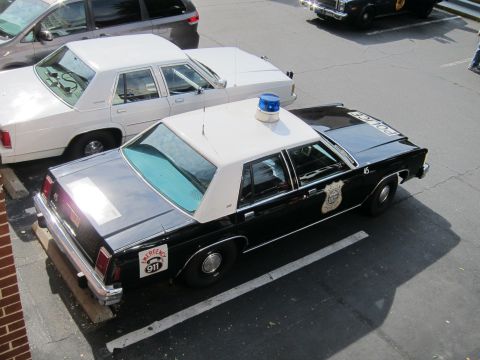 1991 Ford LTD Crown Victoria - overhead view of a black-and-white Ford LTD police sedan with a blue roof beacon and badge decal parked in a lot.