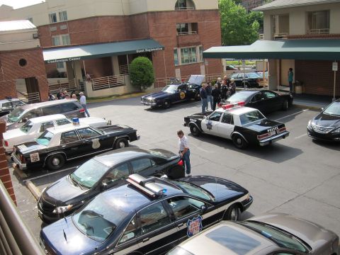 1991 Ford LTD Crown Victoria - elevated view of several police cruisers parked in front of a brick hotel conference center as people walk among the cars.