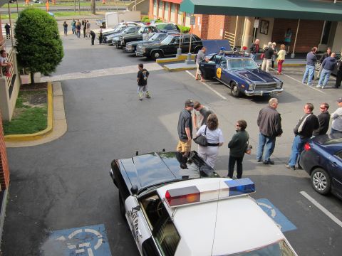 1991 Ford LTD Crown Victoria - elevated view of a police car show with several vintage and modern cruisers parked in a motel lot as a crowd of people looks on.