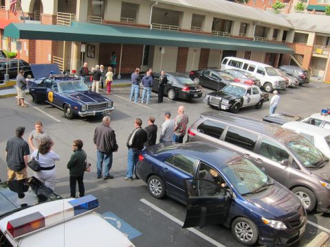1991 Ford LTD Crown Victoria - elevated view of a crowd gathered around vintage and modern police cars, including a blue cruiser and a Ford SUV, in a motel parking lot.