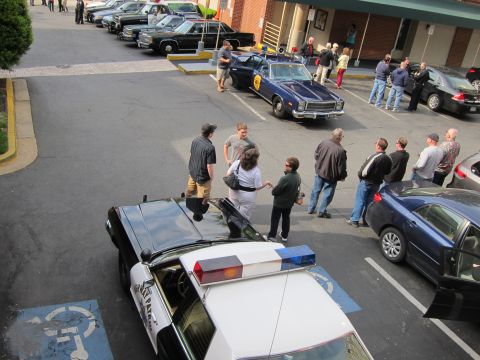 1991 Ford LTD Crown Victoria - Aerial view of vintage police cars, including a black-and-white cruiser, parked outside a motel as a crowd of people mingles in the lot.
