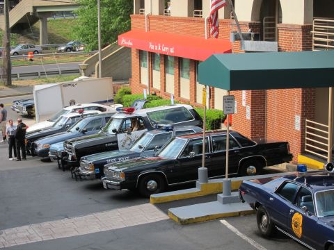 1991 Ford LTD Crown Victoria - lineup of classic black-and-white State Police sedans parked along a brick building marked for pizza carry-out, with an American flag overh…