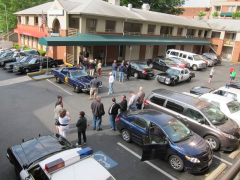 1991 Ford LTD Crown Victoria - Elevated view of a mix of vintage and modern police vehicles parked outside a motel while groups of people stand around the lot.