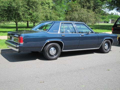 1991 Ford LTD Crown Victoria - Rear view of a dark blue full-size four-door sedan with steel wheels parked on pavement, another cruiser visible at right.
