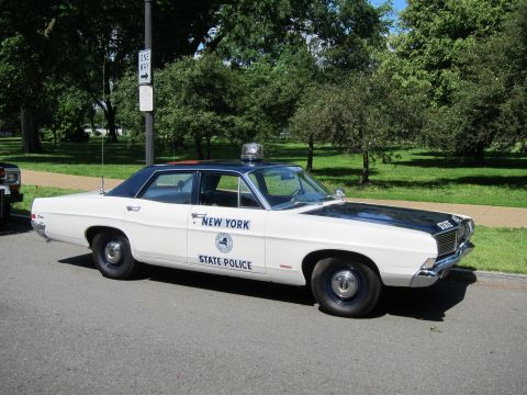 1991 Ford LTD Crown Victoria - white New York State Police sedan with black top and roof beacon parked at an angle on pavement near a parking sign.