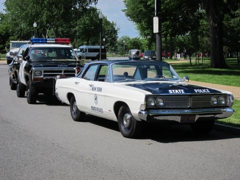 1991 Ford LTD Crown Victoria - white New York State Police sedan parked in line behind a black-and-white Highway Patrol Ramcharger on a tree-lined roadside.