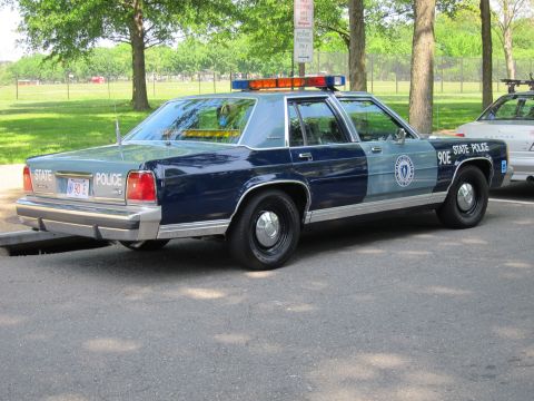 1991 Ford LTD Crown Victoria - Rear view of a two-tone blue and silver State Police sedan with roof light bar, parked beside another cruiser in a grassy lot.